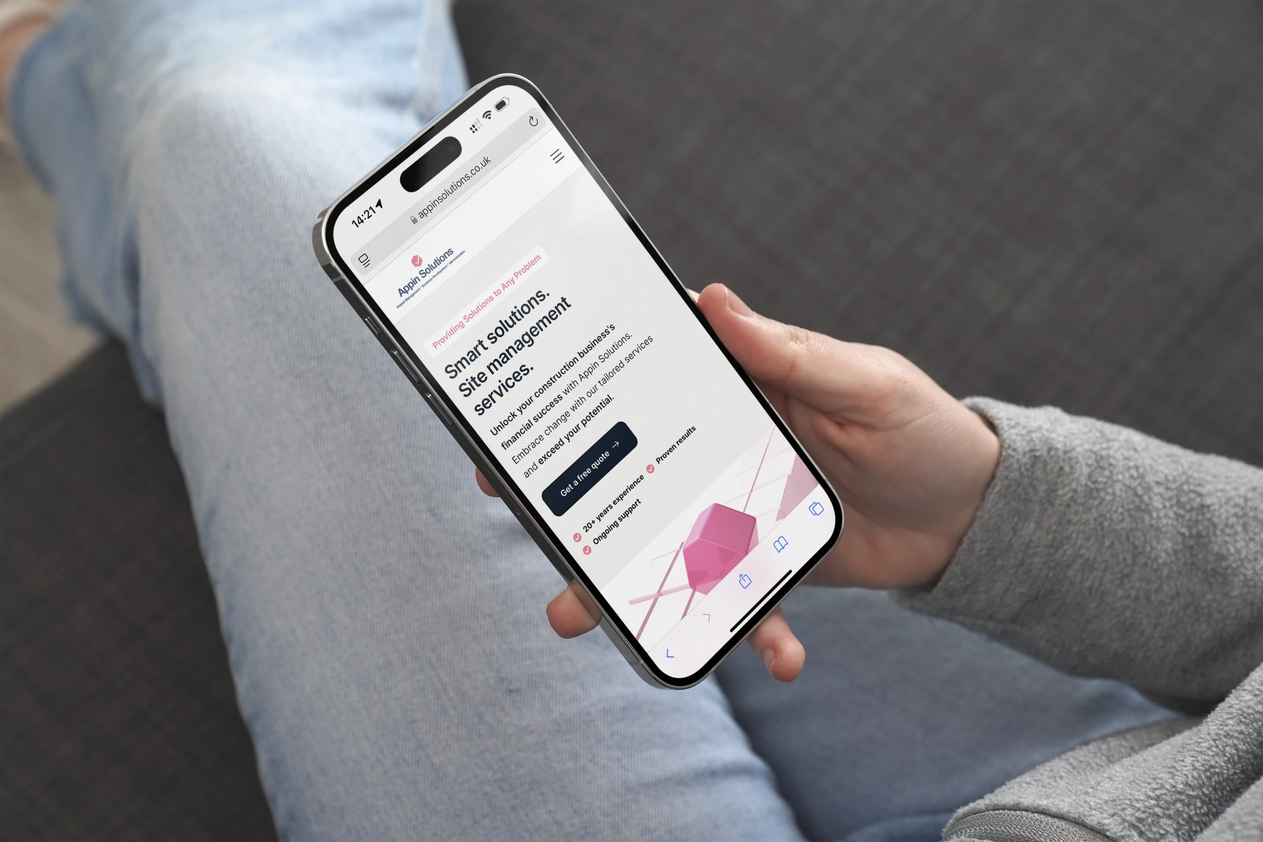 Person holding a smartphone displaying a website about smart solutions for self-management. The person is seated with legs crossed and wearing blue jeans and a gray sweater.